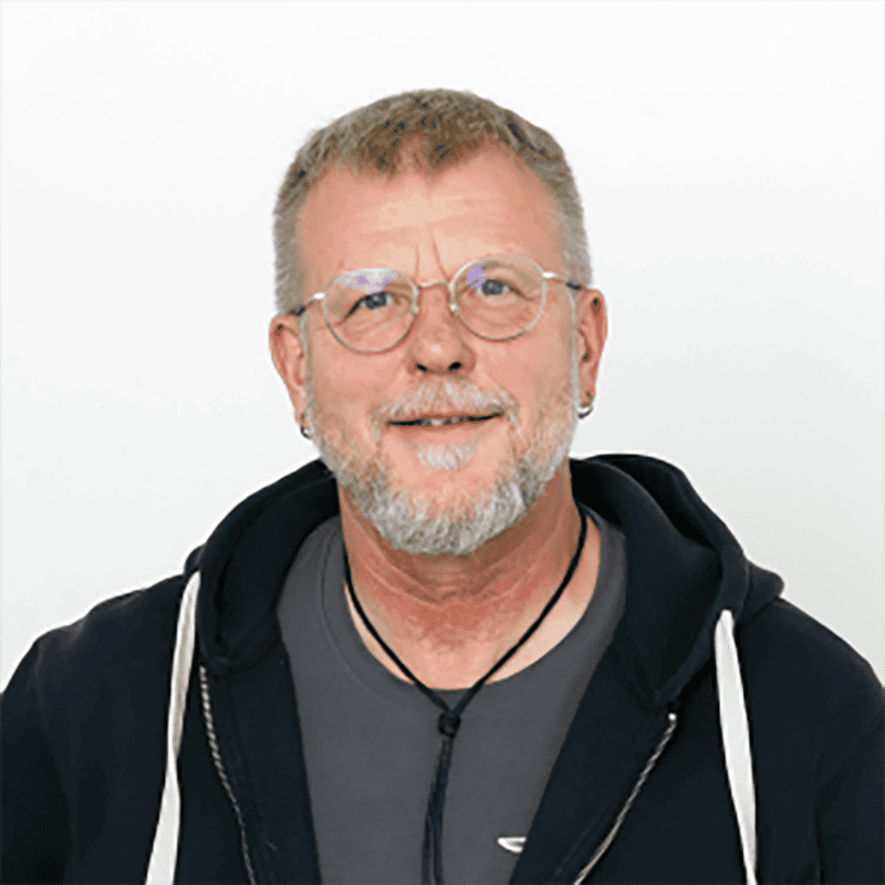 Mann mit Brille und grauem Bart, freundlich lächelnd, in schwarzer Kapuzenjacke und casual Kleidung.